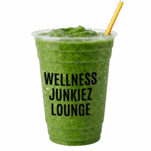 Green spinach and ginger The Detox Dealer smoothie in a clear cup with Wellness Junkiez Lounge logo, yellow straw, fresh spinach leaves, sliced ginger root, and microgreens on a white background.