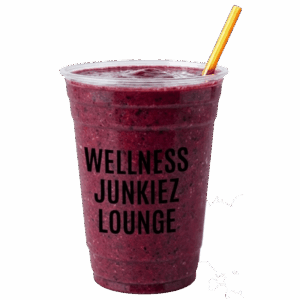 Slim Dealer mixed berry smoothie in a clear cup with Wellness Junkiez Lounge logo, yellow straw, and fresh blueberries, raspberries, strawberries, and blackberries on a white background.